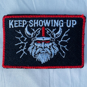 May include: Embroidered patch with the text "KEEP SHOWING UP" above a detailed Viking warrior design. The patch features a black background with a red border and white and red accents.