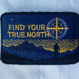 May include: Embroidered patch with the text "FIND YOUR TRUE NORTH" in gold. The patch features a compass rose, a silhouette of a person, and a mountain range in shades of blue and gold against a navy blue background.