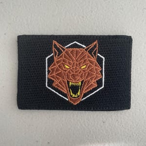 May include: A rectangular black patch featuring a geometric wolf head design. The wolf is brown with yellow eyes and a white outline, set within a white hexagonal frame. The patch is likely made of fabric.