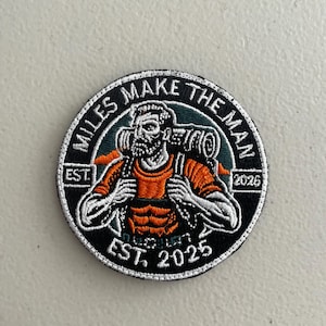 May include: Circular patch with a hiker graphic, backpack, and the text "MILES MAKE THE MAN." The patch features an orange and black color scheme with the text "EST. 2025" and "2026."
