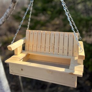 May include: A light-colored wooden bird feeder designed to resemble a miniature porch swing. It features a slatted backrest, two armrests, and a rectangular base with a mesh floor. The feeder is suspended by silver metal chains attached to the sides.