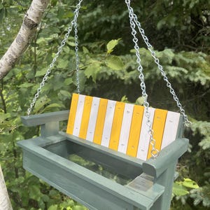 May include: A hanging bird feeder shaped like a porch swing with a sage green frame and a backrest featuring alternating vertical yellow and white wooden slats. The feeder includes a mesh base for drainage and is suspended by silver metal chains.