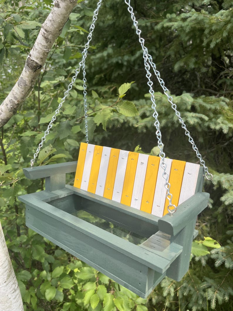 May include: A hanging bird feeder shaped like a porch swing with a sage green frame and a backrest featuring alternating vertical yellow and white wooden slats. The feeder includes a mesh base for drainage and is suspended by silver metal chains.