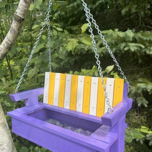 May include: A wooden bird feeder shaped like a porch swing, featuring a purple frame and a backrest with alternating vertical yellow and white slats. The feeder is suspended by silver metal chains attached to the armrests on both sides.
