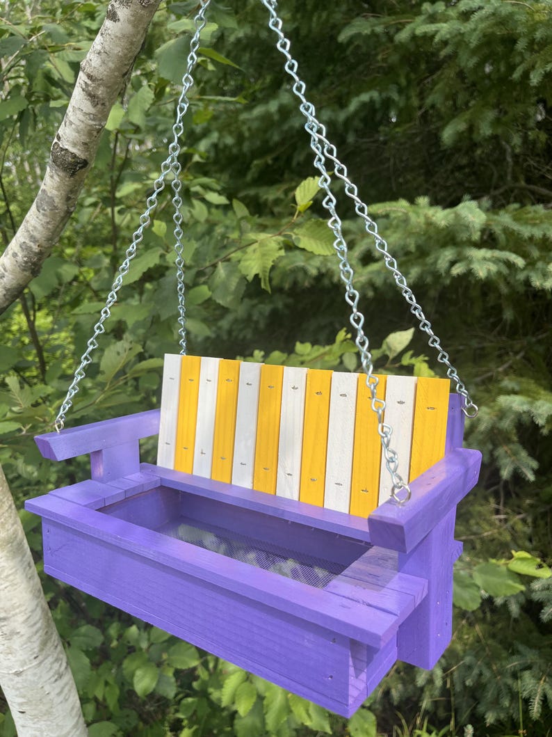 May include: A wooden bird feeder shaped like a porch swing, featuring a purple frame and a backrest with alternating vertical yellow and white slats. The feeder is suspended by silver metal chains attached to the armrests on both sides.