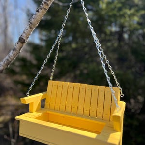 May include: A bright yellow wooden bird feeder designed to resemble a miniature porch swing. It features a slatted backrest, two armrests, and a rectangular base tray for birdseed, suspended by silver metal chains from a single hook.