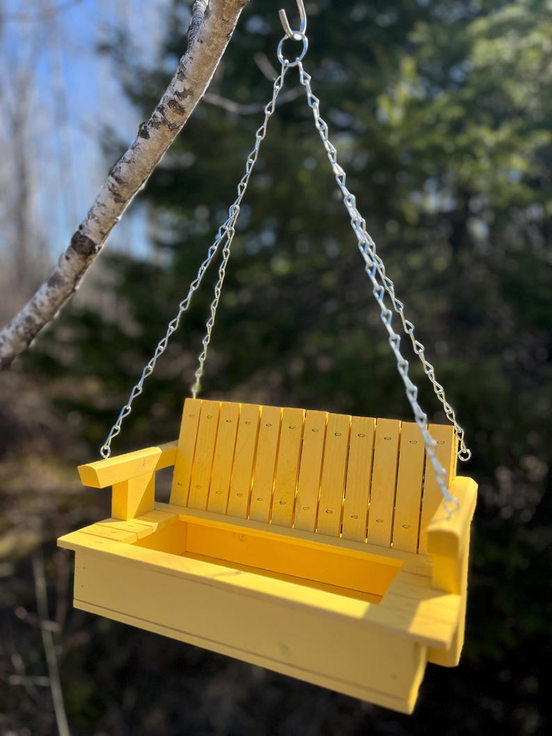 May include: A bright yellow wooden bird feeder designed to resemble a miniature porch swing. It features a slatted backrest, two armrests, and a rectangular base tray for birdseed, suspended by silver metal chains from a single hook.