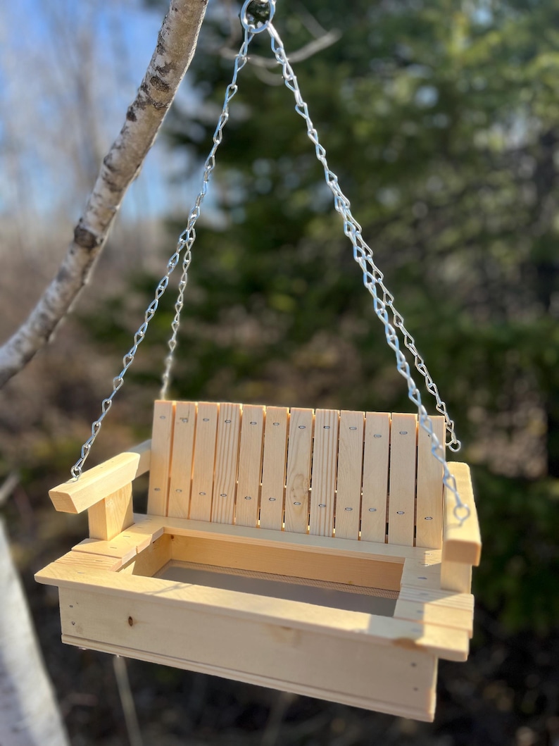 May include: A light-colored wooden bird feeder designed to resemble a miniature porch swing. It features a slatted backrest, two armrests, and a rectangular base with a mesh floor. The feeder is suspended by silver metal chains attached to the sides.