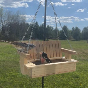 May include: A light-colored wooden bird feeder shaped like a porch swing, suspended by silver metal chains from a central black pole. Two small birds are perched on the wooden slats of the feeder, which features a backrest and armrests.