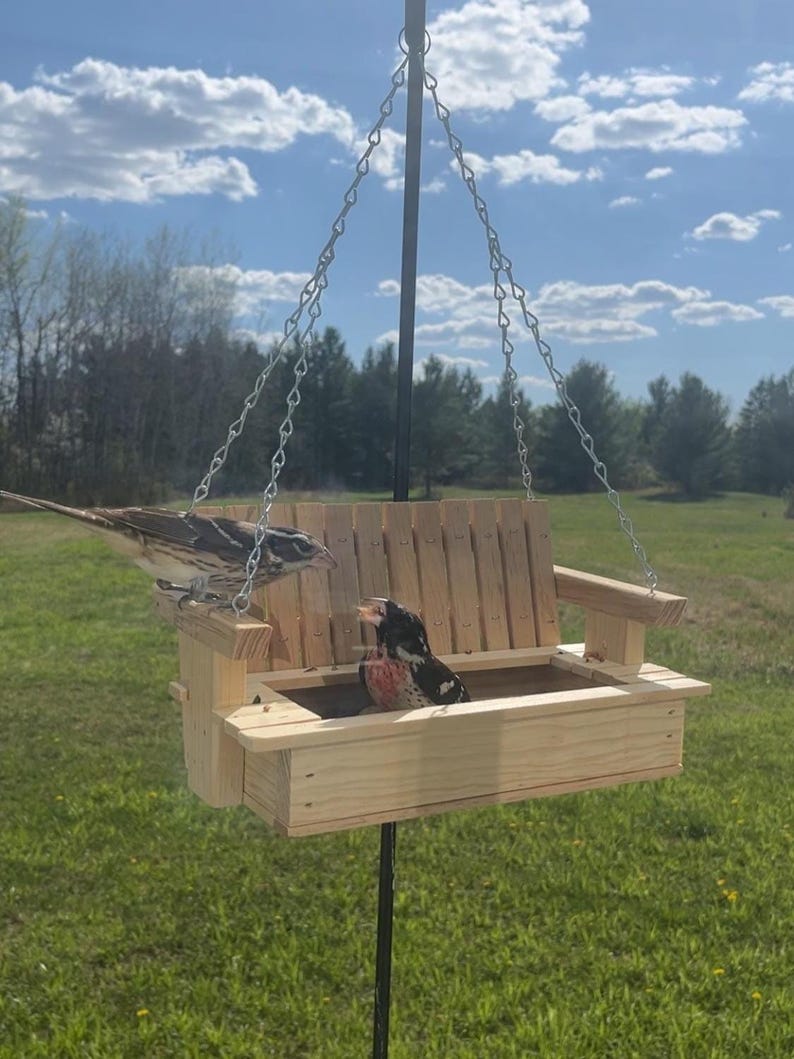May include: A light-colored wooden bird feeder shaped like a porch swing, suspended by silver metal chains from a central black pole. Two small birds are perched on the wooden slats of the feeder, which features a backrest and armrests.