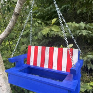 May include: A hanging bird feeder shaped like a bench, featuring a bright blue wooden frame and a backrest with alternating vertical red and white slats. The structure is suspended by silver metal chains attached to the armrests.