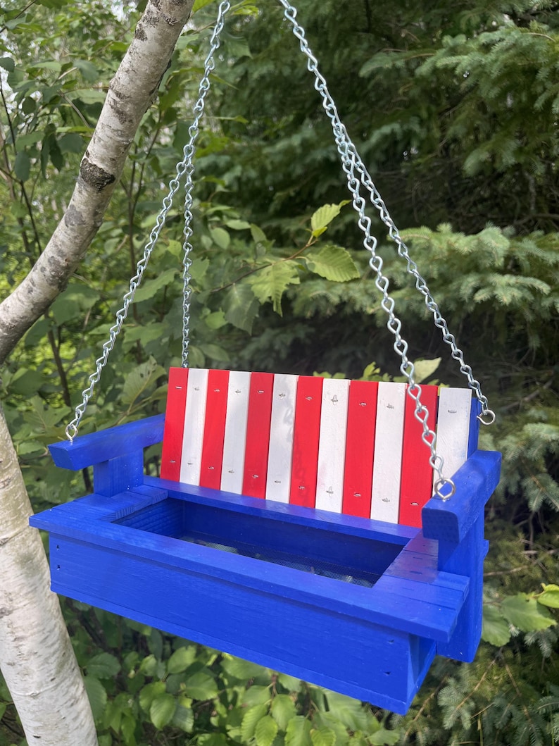 May include: A hanging bird feeder shaped like a bench, featuring a bright blue wooden frame and a backrest with alternating vertical red and white slats. The structure is suspended by silver metal chains attached to the armrests.