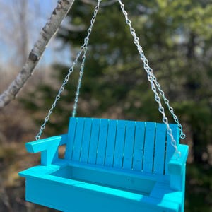 May include: A bright turquoise wooden bench planter suspended by silver metal chains. The bench features a slatted backrest, flat armrests, and a rectangular open compartment designed for holding items, hanging from a single metal ring.