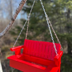 May include: A bright red wooden porch swing bird feeder featuring a slatted backrest, flat armrests, and a rectangular base tray for seeds. The feeder is suspended by four silver-toned metal chains that connect to a single top hanging ring.