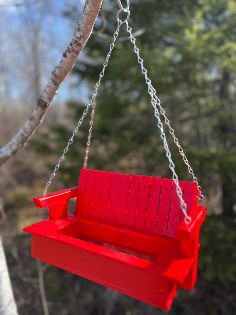May include: A bright red wooden porch swing bird feeder featuring a slatted backrest, flat armrests, and a rectangular base tray for seeds. The feeder is suspended by four silver-toned metal chains that connect to a single top hanging ring.