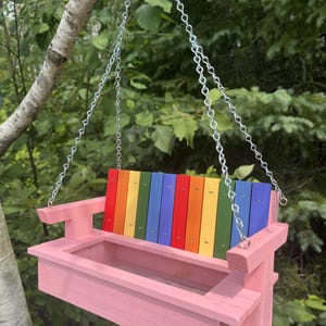 May include: A pink wooden bird feeder shaped like a porch swing, featuring a backrest made of vertical slats painted in a rainbow gradient of red, orange, yellow, green, blue, and purple. The feeder hangs from a silver metal chain.