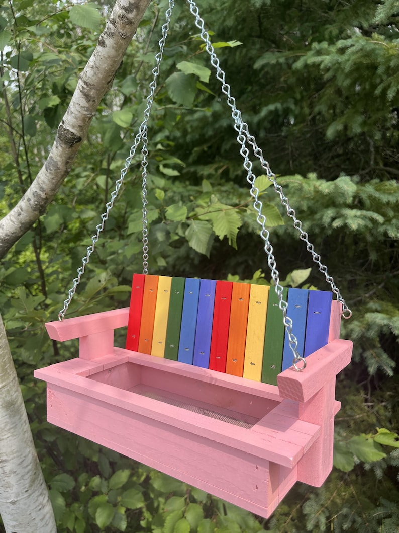 May include: A pink wooden bird feeder shaped like a porch swing, featuring a backrest made of vertical slats painted in a rainbow gradient of red, orange, yellow, green, blue, and purple. The feeder hangs from a silver metal chain.