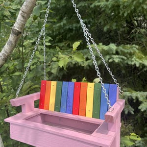 May include: A hanging bird feeder constructed from wood, featuring a light pink base and armrests. The backrest consists of vertical slats painted in a rainbow sequence of red, orange, yellow, green, and blue. It is suspended by silver metal chains.