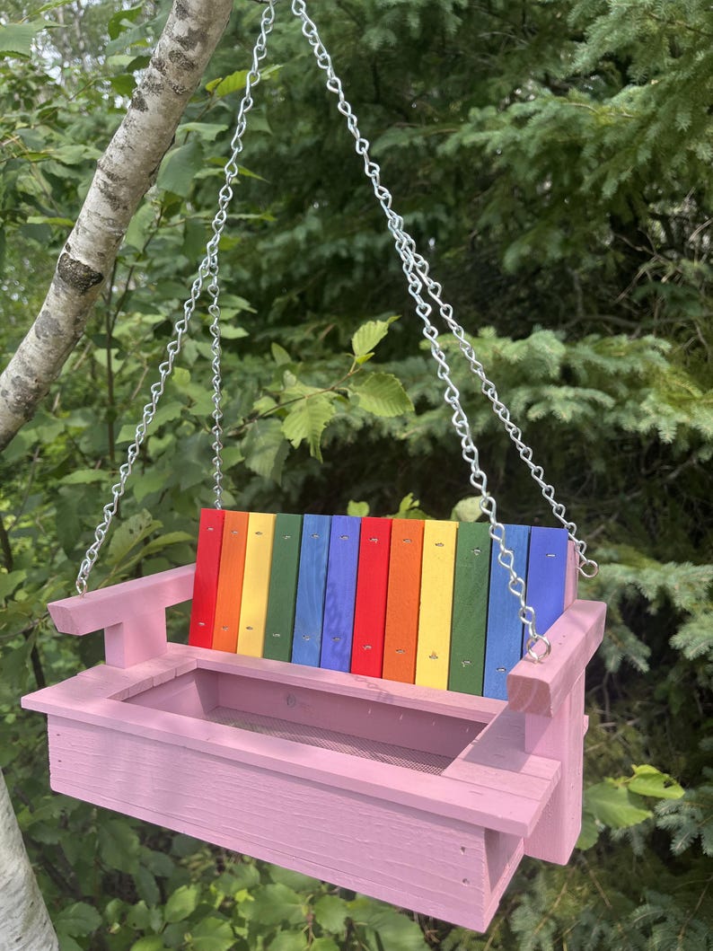 May include: A hanging bird feeder constructed from wood, featuring a light pink base and armrests. The backrest consists of vertical slats painted in a rainbow sequence of red, orange, yellow, green, and blue. It is suspended by silver metal chains.