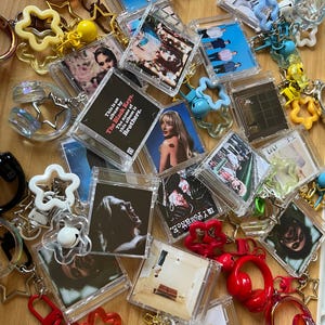May include: A collection of miniature square keychains featuring various album cover art in clear plastic cases. The keychains are attached to colorful star-shaped charms, small bells, and miniature headphone charms in red, yellow, blue, and white.