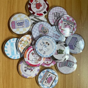 May include: A selection of clear, circular acrylic badges with diverse, vibrant designs. Text on the badges includes "COZY GAMER," "TOO MANY THBS," and "Self love club." Other designs feature a goose, a heart, and a retro computer.