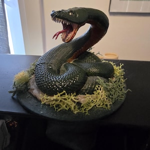 Peut inclure: Une figurine de serpent vert avec une langue rouge, la bouche ouverte et des crocs. Le serpent est enroulé autour d'une base avec de la mousse verte et de la terre brune.