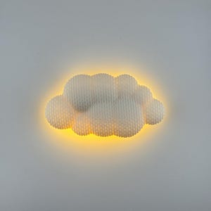 Könnte beinhalten: Eine weiße, wolkenförmige Wandleuchte mit strukturierter Oberfläche. Das Licht strahlt ein warmes gelbes Leuchten von hinter der Wolke aus und erzeugt einen weichen, umgebenden Effekt. Die Leuchte ist an einer schlichten, hellgrauen Wand befestigt.