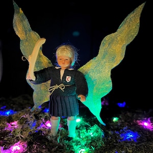 Peut inclure: Une poupée fée miniature aux cheveux blonds, vêtue d'un uniforme scolaire bleu foncé et de bottes blanches, tenant un arc en bois. La poupée a de grandes ailes translucides avec des accents dorés et verts. La fée se tient sur de la mousse, éclairée par des guirlandes lumineuses colorées.