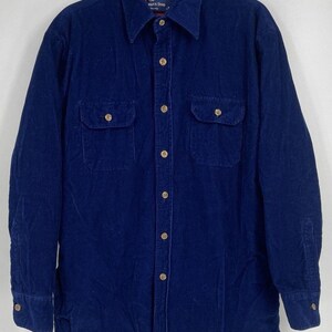 May include: A long-sleeve navy blue corduroy shirt featuring a button-down front, a pointed collar, and two chest pockets with buttoned flaps. The interior collar lining is red, and the shirt includes a label reading The Men's Shop.