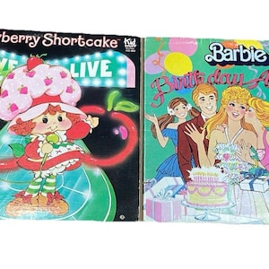 Könnte beinhalten: Zwei Vintage-Vinyl-Schallplattenalben. Eines zeigt Erdbeerkuchen mit dem Text "LIVE" und das andere ist ein "Geburtstagsalbum" von Barbie mit Illustrationen von Barbie und ihren Freunden. Beide Alben haben farbenfrohe Designs.