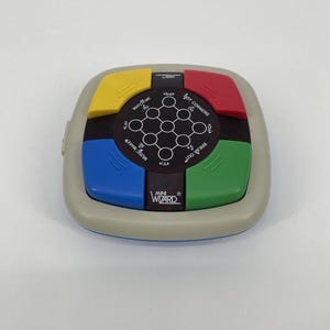 May include: A handheld Mini Wizard electronic game with a gray casing and colorful buttons. The buttons are yellow, red, green, and blue. The game has a black center with white circular patterns and text that reads "MATCH ME", "MUSIC MAKER", "HOT CORNERS", and "BREAK OUT".