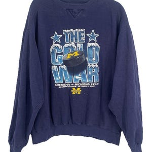 May include: Navy blue sweatshirt with the text "THE COLD WAR" in a stylized font, featuring a hockey puck graphic. The sweatshirt commemorates the Michigan vs. Michigan State game at Spartan Stadium on October 6, 2001.