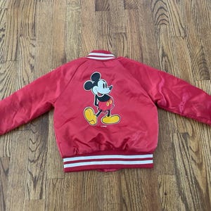 May include: Red satin jacket with white and red striped trim on the collar, cuffs, and hem. The back features a large graphic of Mickey Mouse in his classic pose. A vintage-style jacket.