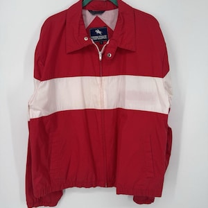Puede incluir: Chaqueta roja y blanca con cremallera, cuello y dos bolsillos delanteros. La chaqueta tiene una franja horizontal blanca en el pecho y el texto "STEEPLECHASE" en la etiqueta. Colgada de una percha verde.