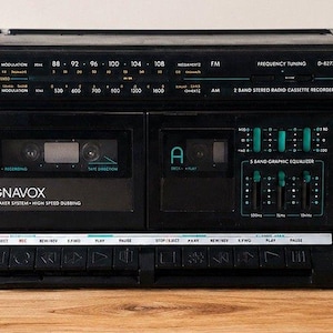 Pode incluir: Um boombox Magnavox preto vintage com dois alto-falantes e um toca-fitas. O boombox possui um sintonizador de rádio, equalizador gráfico e controles de volume. Os alto-falantes são rotulados como "2 Way Speaker System".