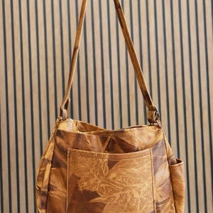 May include: A brown leather shoulder bag with a large front pocket and a side pocket. The bag has long straps and a textured, patterned design. The bag is held up by a hand.