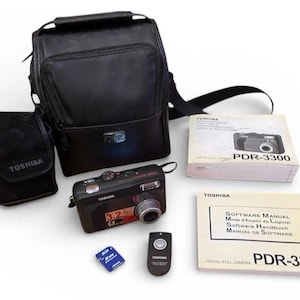 May include: A black Toshiba PDR-3300 digital camera kit. The kit includes a camera, a carrying case, a smaller case, a remote control, a memory card, and two manuals. The camera has a 3.2x zoom lens.