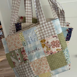 May include: A patchwork tote bag with a light blue and white gingham pattern. The bag features a variety of floral and patterned fabric squares in pastel colors. The name "Brooke" is embroidered on the front.