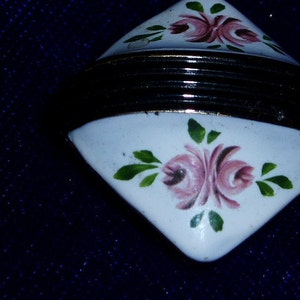 May include: A white ceramic trinket box with a black and gold trim. The box is decorated with pink and green floral designs.