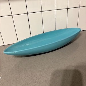 May include: A long, oval-shaped, turquoise-colored ceramic serving dish. The dish has a smooth, glossy finish and is set on a gray countertop. The background includes white tiles and an electrical outlet.