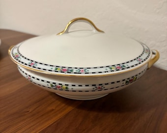 May include: A white ceramic serving bowl with a lid. The bowl has a gold handle and gold accents. A black and white geometric pattern with floral accents decorates the rim. The bowl is on a wooden surface.