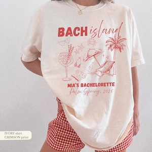 May include: Ivory-colored t-shirt with a crimson graphic design. The design includes the words "BACH island" and "MIA'S BACHELORETTE Palm Spring, 2025" along with illustrations of a cocktail, sunglasses, and a beach scene.