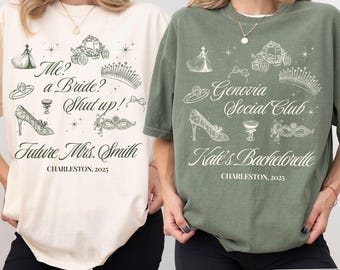May include: Two oversized t-shirts with a white and green color scheme. The ivory shirt has a black print that says "Me? a Bride? Shut up! Future Mrs. Smith CHARLESTON, 2025". The moss green shirt has a black print that says "Genovia Social Club Kate's Bachelorette CHARLESTON, 2025". Both shirts feature a princess crown, a carriage, a glass slipper, a mask, and a glass of champagne.
