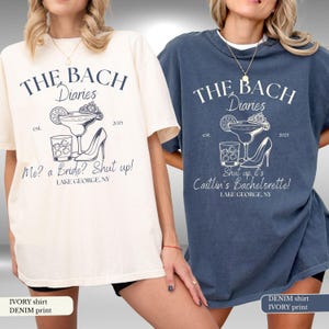 May include: Two t-shirts, one ivory and one denim, with a graphic print. The ivory shirt reads "THE BACH Diaries" with a cocktail, high heel, and text "Me? a Bride? Shut up! LAKE GEORGE, NY." The denim shirt says "Shut up, it's Caitlin's Bachelorette!"