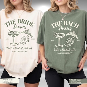 May include: Two t-shirts, one cream and one khaki green, featuring a graphic design. The design includes the text "THE BRIDE Diaries" and "THE BACH Diaries" with a cocktail glass, high heels, and a tiara. The shirts also have the text "Me? a Bride? Shut up!" and "Kate's Bachelorette".