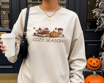May include: Sand-colored sweatshirt with a fall-themed graphic. The graphic includes books, a pumpkin, a coffee cup, and the words "COZY SEASON." The sweatshirt is a neutral color and has a crew neck.