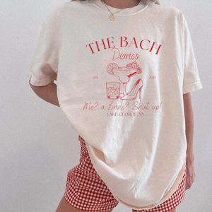 May include: Cream-colored t-shirt with a red graphic that reads "THE BACH Diaries" with a cocktail, high heel, and text "Me? a Bride? Shut up!" The shirt is a relaxed fit.