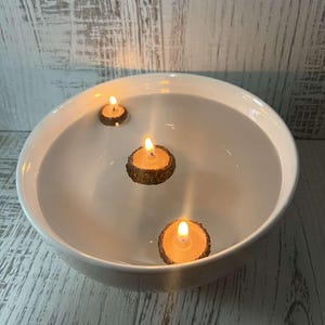May include: Three small, round, wooden candles are floating in a white bowl filled with water. The candles are lit and the flames are visible.