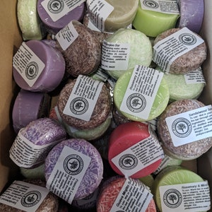May include: A box filled with various round, colorful, solid shampoo bars. The bars are wrapped in clear plastic and have labels with different scents and ingredients, such as Guava Mojito, Rosemary & Mint, and Sandalwood.