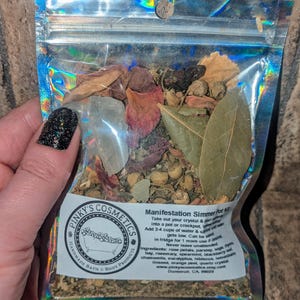Può includere: Un sacchetto trasparente e iridescente pieno di erbe secche, foglie e cristalli. L'etichetta recita "Manifestation Simmer Pot Kit" ed elenca ingredienti come petali di rosa, salvia ed eucalipto. È visibile anche il marchio "Pinky's Cosmetics".